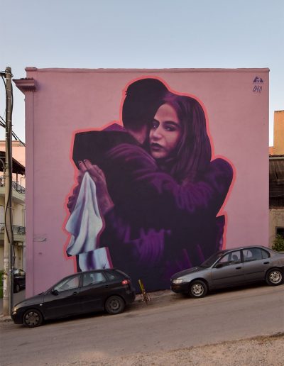 Mural by emic in Patras, Greece featuring two figures hugging with the female gazing out to the viewer as she holds a white cloth to represent peace